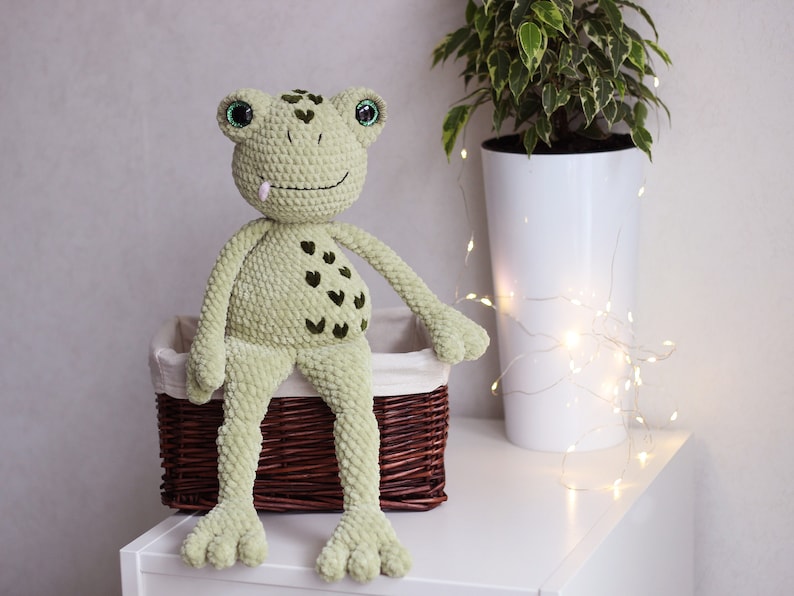 May include: A green crocheted frog with a pink tongue sitting in a brown wicker basket. The frog has green eyes and a pattern of dark green hearts on its belly.