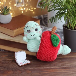 May include: A green crocheted worm wearing glasses sits next to a red crocheted apple with a green leaf. The worm and apple are on a wooden surface with a stack of books and a potted plant in the background.