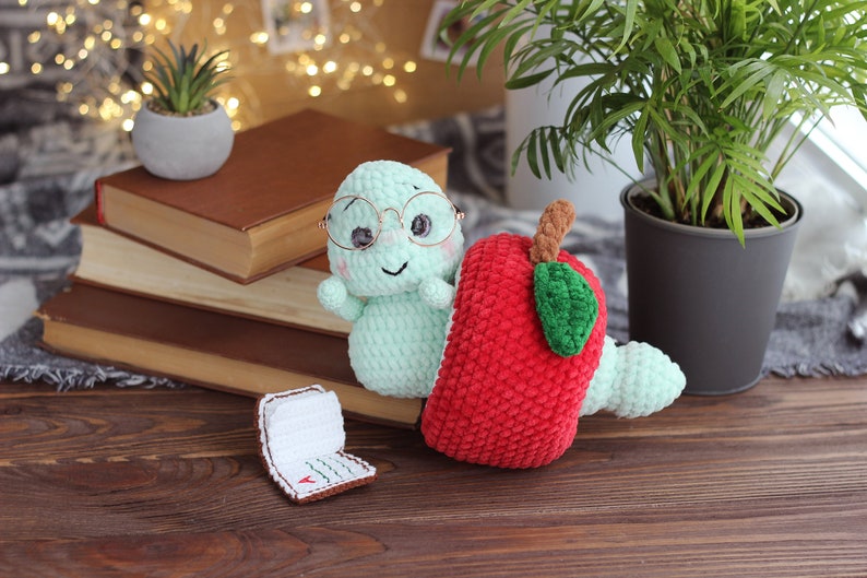 May include: A green crocheted worm wearing glasses sits next to a red crocheted apple with a green leaf. The worm and apple are on a wooden surface with a stack of books and a potted plant in the background.