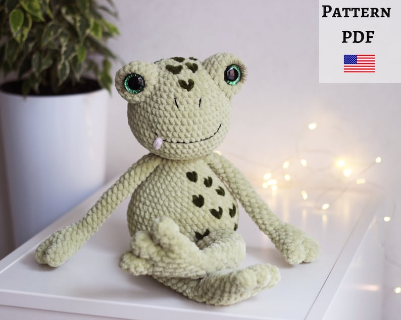 May include: Green crocheted frog amigurumi toy with black heart details and green eyes. The frog is sitting on a white surface.
