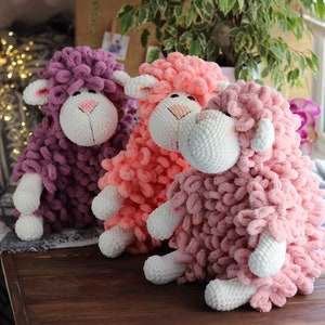 May include: Three crocheted stuffed sheep toys. One is purple, one is pink, and one is a light peach color. They are all sitting on a wooden table.