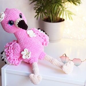 May include: Pink crocheted flamingo plush toy with white flowers and blue eyes. The flamingo is sitting on a white surface with a pair of heart-shaped sunglasses.