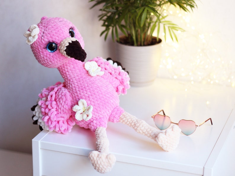 May include: Pink crocheted flamingo plush toy with white flowers and blue eyes. The flamingo is sitting on a white surface with a pair of heart-shaped sunglasses.