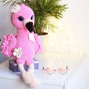 May include: Pink crocheted flamingo plush toy with white flowers and blue eyes. The flamingo is sitting on a white surface with a box and heart-shaped sunglasses.