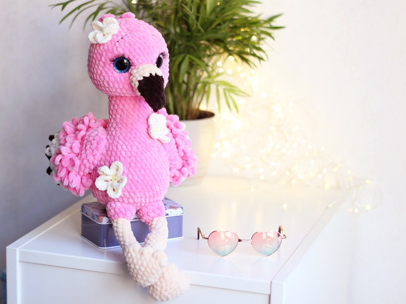 May include: Pink crocheted flamingo plush toy with white flowers and blue eyes. The flamingo is sitting on a white surface with a box and heart-shaped sunglasses.