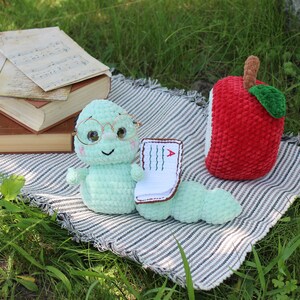 May include: A green crocheted worm wearing glasses and holding a small book with a red 'A' on it. The worm is sitting on a striped blanket with a red crocheted apple beside it.