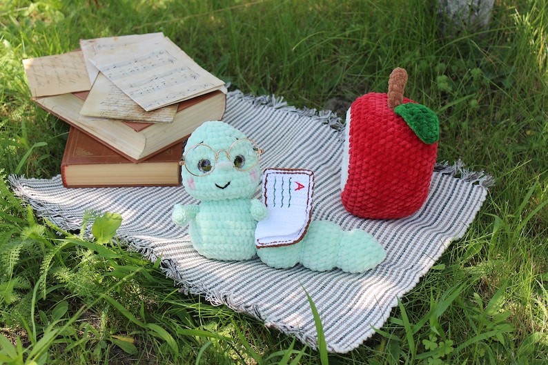 May include: A green crocheted worm wearing glasses and holding a small book with a red 'A' on it. The worm is sitting on a striped blanket with a red crocheted apple beside it.