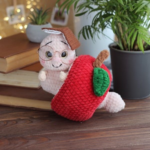 May include: A crocheted worm wearing glasses and a brown hat, peeking out of a red crocheted apple.