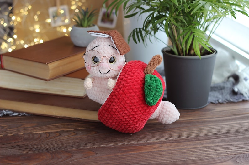 May include: A crocheted worm wearing glasses and a brown hat, peeking out of a red crocheted apple.