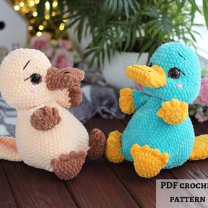 May include: Two handmade crochet platypus toys. One is beige and brown, the other is teal and yellow. Both have large eyes, a beak, and webbed feet. The toys are sitting on a wooden surface. The image includes the text "PDF CROCHET PATTERN" and a small American flag.