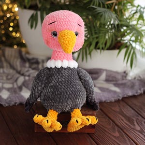 May include: A handmade plush vulture toy with a pink head, yellow beak, and black eyes. The vulture has a gray body, white collar, and yellow feet. The toy is sitting on a wooden surface.