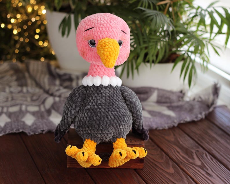 May include: A handmade plush vulture toy with a pink head, yellow beak, and black eyes. The vulture has a gray body, white collar, and yellow feet. The toy is sitting on a wooden surface.