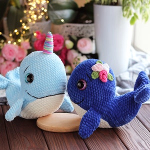 May include: Two crocheted whale plushies, one light blue with a rainbow horn and the other dark blue with pink flowers on its head.
