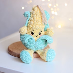 May include: A crocheted corn on the cob plush toy with a smiling face. The corn is yellow and the husk is light blue. The toy is sitting on a wooden surface.