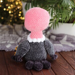 May include: A handmade plush vulture toy. The vulture has a pink head and neck, a white and gray collar, and a gray body. The feet are dark gray. The toy is made of soft, textured fabric and is sitting on a wooden surface.