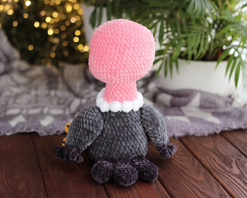 May include: A handmade plush vulture toy. The vulture has a pink head and neck, a white and gray collar, and a gray body. The feet are dark gray. The toy is made of soft, textured fabric and is sitting on a wooden surface.