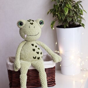 May include: A green crocheted frog plush toy with green eyes and a pink tongue, sitting on a brown wicker basket.