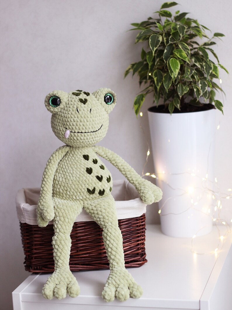 May include: A green crocheted frog plush toy with green eyes and a pink tongue, sitting on a brown wicker basket.