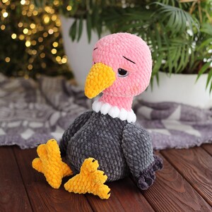 May include: A handmade plush vulture toy with a pink head, yellow beak, and gray body. The vulture has white collar details and yellow feet. The toy is made of soft, textured yarn and sits on a wooden surface.