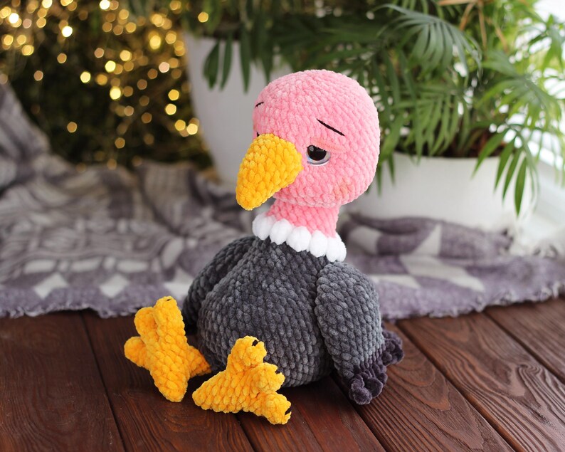 May include: A handmade plush vulture toy with a pink head, yellow beak, and gray body. The vulture has white collar details and yellow feet. The toy is made of soft, textured yarn and sits on a wooden surface.