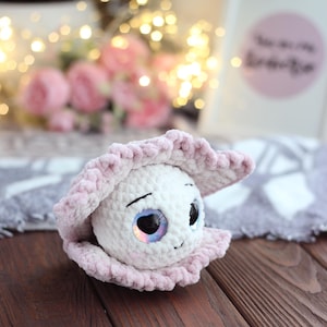 Puede incluir: Una perla blanca hecha a mano, tejida a ganchillo, con ojos iridiscentes y una pequeña sonrisa, metida en una concha rosa claro tejida a ganchillo. El peluche está sobre un fondo borroso de flores rosas y luces de hadas, sobre una superficie de madera oscura.