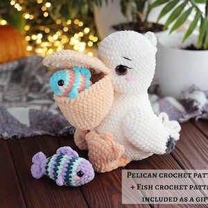May include: A white and peach crochet pelican plush toy with a large beak holding a small blue and white striped crochet fish. A second purple, green, and white striped crochet fish sits nearby. Text reads: PELICAN CROCHET PATTERN + FISH CROCHET PATTERN INCLUDED AS A GIFT.