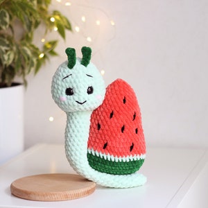 Può includere: Una lumaca verde all'uncinetto con un viso sorridente e un guscio di anguria. L'anguria è rossa con semi neri e una buccia verde.