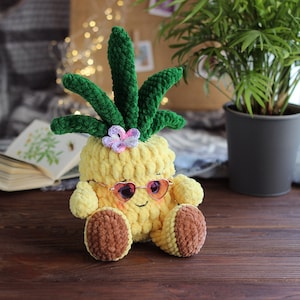 Könnte beinhalten: Ein gehäkeltes Ananas-Spielzeug, das eine rosa Sonnenbrille trägt. Die Ananas ist gelb mit grünen Blättern und einer rosa Blume oben. Das Spielzeug sitzt auf einer Holzoberfläche.