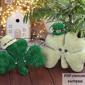 Può includere: Due soffici trifogli verdi con facce sorridenti e piccoli cappelli. Un trifoglio indossa un cappello color crema con una fascia marrone, mentre l'altro indossa un cappello verde. Lo sfondo include una casa bianca con luci e una pianta.