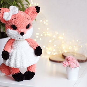 May include: A crocheted plush fox toy with a white dress and a pink bow. The fox has black eyes and a black nose. The toy is sitting on a white surface.