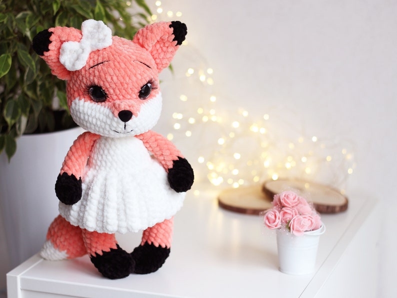 May include: A crocheted plush fox toy with a white dress and a pink bow. The fox has black eyes and a black nose. The toy is sitting on a white surface.