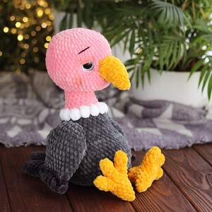 May include: A handmade plush vulture toy with a pink head, gray body, and yellow beak and feet. The vulture has a white ruffled collar and a slightly sad expression. The toy is made of soft, textured yarn and sits on a wooden surface.