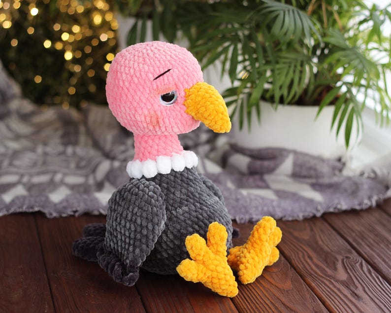 May include: A handmade plush vulture toy with a pink head, gray body, and yellow beak and feet. The vulture has a white ruffled collar and a slightly sad expression. The toy is made of soft, textured yarn and sits on a wooden surface.