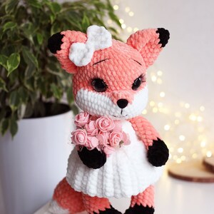 May include: A crocheted fox plush toy wearing a white dress and holding a bouquet of pink roses. The fox has a pink body, black ears and feet, and a white bow on its head.