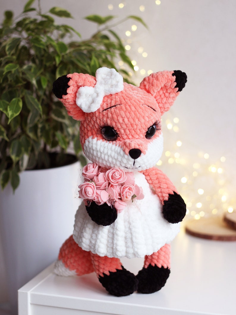 May include: A crocheted fox plush toy wearing a white dress and holding a bouquet of pink roses. The fox has a pink body, black ears and feet, and a white bow on its head.