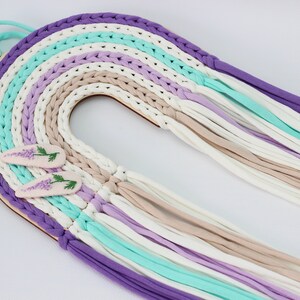 Puede incluir: Un tapiz de macramé de colores arcoíris con una base de madera y detalles de flores moradas.