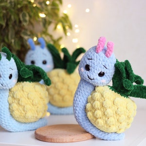 Può includere: Tre peluche di lumaca all'uncinetto con gusci di ananas. Le lumache sono blu con ananas gialli e foglie verdi. Una lumaca ha antenne rosa.