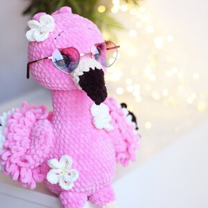 May include: Pink crocheted flamingo toy wearing heart-shaped sunglasses with white crocheted flowers on its head.