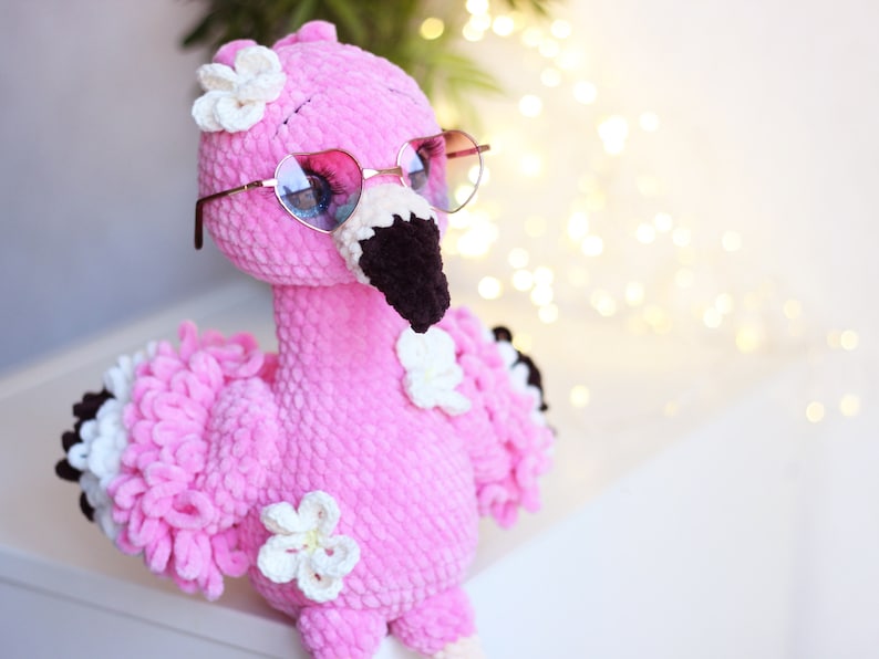 May include: Pink crocheted flamingo toy wearing heart-shaped sunglasses with white crocheted flowers on its head.