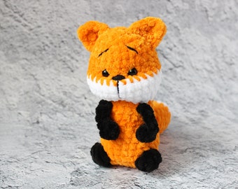 May include: A crocheted orange fox plush toy with white and black accents. The fox has a white muzzle and black eyes. The toy is sitting on a gray textured surface.