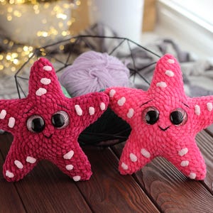 Könnte beinhalten: Zwei Plüsch-Seestern-Spielzeuge, eines in tiefem Pink und eines in hellem Pink, jeweils mit weißen Tupfen und großen, ausdrucksstarken Augen. Die Spielzeuge haben ein lächelndes Gesicht und sind aus einem weichen, strukturierten Material gefertigt. Sie liegen auf einer Holzoberfläche.