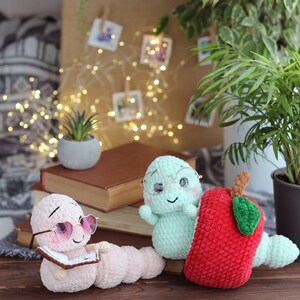 May include: Two crocheted worm plushies, one wearing pink heart-shaped sunglasses and reading a book, the other wearing round glasses and sitting next to a red crocheted apple.