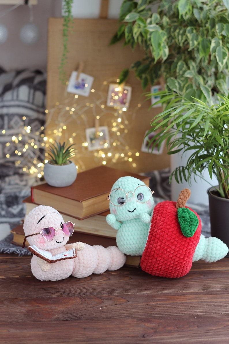 May include: Two crocheted worm plushies, one wearing pink heart-shaped sunglasses and reading a book, the other wearing round glasses and sitting next to a red crocheted apple.