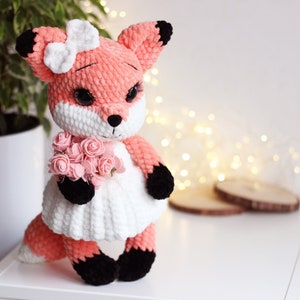May include: A crocheted fox plush toy wearing a white dress and holding a bouquet of pink flowers. The fox has black eyes, a black nose, and black paws.