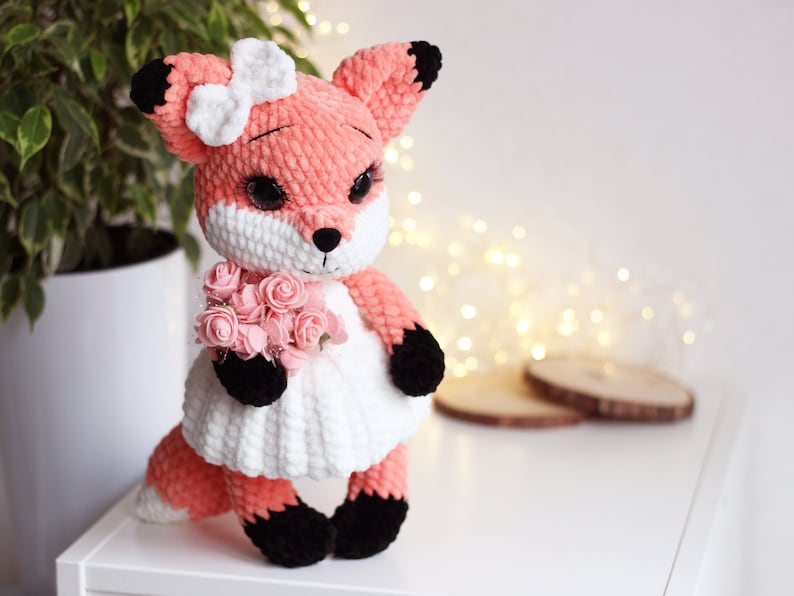 May include: A crocheted fox plush toy wearing a white dress and holding a bouquet of pink flowers. The fox has black eyes, a black nose, and black paws.