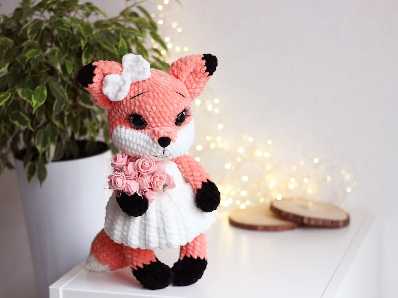 May include: A crocheted fox plush toy wearing a white dress and holding a bouquet of pink flowers. The fox has black eyes, a pink nose, and black ears.