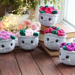 May include: Five crocheted flower pots with happy faces. Each pot is filled with colorful crocheted flowers. The pots are arranged on a wooden surface.