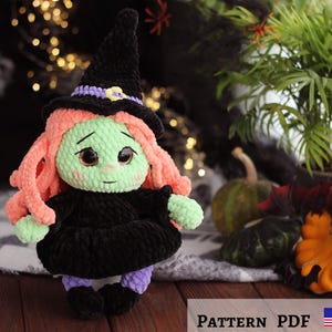 Puede incluir: Una muñeca de bruja de crochet con piel verde, cabello rosa y un sombrero de bruja negro. La muñeca lleva un vestido negro y leggings morados. La muñeca está sentada sobre una superficie marrón con una planta verde en el fondo.