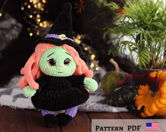 May include: A crocheted witch doll with green skin, pink hair, and a black witch's hat. The doll is wearing a black dress and purple leggings. The doll is sitting on a brown surface with a green plant in the background.