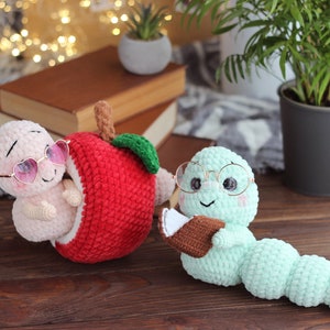 May include: Two crocheted worm plushies, one wearing pink heart-shaped sunglasses and sitting inside a red crocheted apple, the other wearing round glasses and holding a brown crocheted book.
